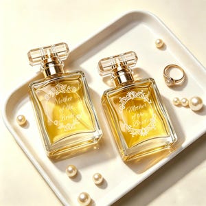 Puede incluir: Dos frascos de perfume rectangulares con detalles dorados y tapones transparentes facetados. Los frascos contienen un líquido dorado y llevan la inscripción "Mother of the Bride". Se encuentran sobre una bandeja blanca, junto a perlas y un anillo de oro.