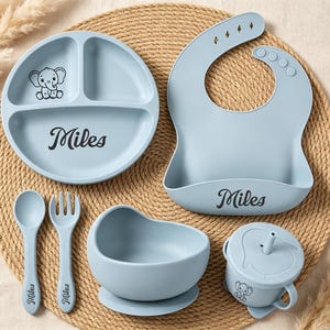 May include: A light blue silicone feeding set featuring a divided plate, a bib, a bowl, a cup with a straw, a spoon, and a fork. The plate, bib, spoon, and fork are personalized with the name Miles. The plate and cup feature a small elephant graphic.
