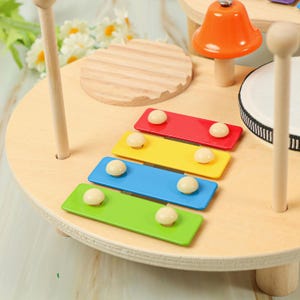Puede incluir: Un juego de instrumentos musicales de juguete de madera. Cuenta con un xil&oacute;fono con teclas rojas, amarillas, azules y verdes. El juego incluye un tambor peque&ntilde;o, una campana y una maza de madera. Los instrumentos est&aacute;n dispuestos sobre una superficie de madera clara.