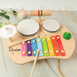 Puede incluir: Juego de instrumentos musicales de juguete de madera con xil&oacute;fono, tambores, platillo y un instrumento de percusi&oacute;n de madera. El xil&oacute;fono tiene barras de colores y los tambores tienen parches blancos. El nombre "Denise" est&aacute; impreso.