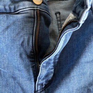 K&ouml;nnte beinhalten: Nahaufnahme einer blauen Jeans mit teilweise ge&ouml;ffnetem Rei&szlig;verschluss. Die Jeans hat einen Knopf mit den Buchstaben "NYDJ". Der Denim hat ein verwaschenes Aussehen.