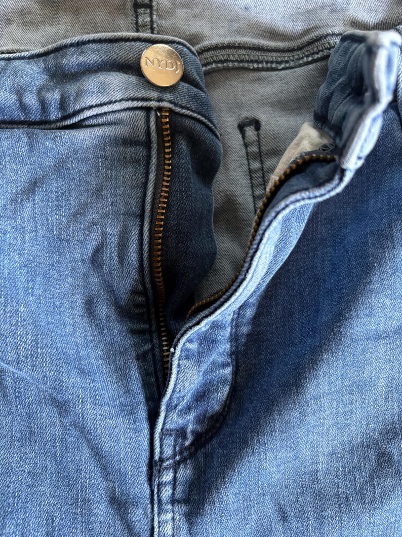 K&ouml;nnte beinhalten: Nahaufnahme einer blauen Jeans mit teilweise ge&ouml;ffnetem Rei&szlig;verschluss. Die Jeans hat einen Knopf mit den Buchstaben "NYDJ". Der Denim hat ein verwaschenes Aussehen.