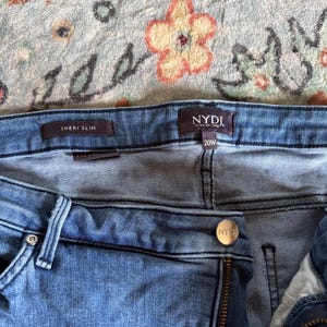 K&ouml;nnte beinhalten: Nahaufnahme einer blauen Jeans mit den sichtbaren Markennamen "NYDJ" und "SHERI SLIM". Die Jeans hat die Gr&ouml;&szlig;e 20W. Der Hintergrund ist ein Stoff mit floralem Muster.