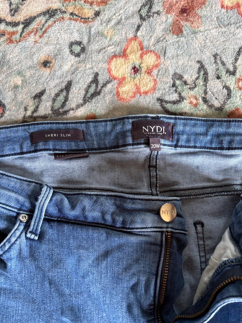 K&ouml;nnte beinhalten: Nahaufnahme einer blauen Jeans mit den sichtbaren Markennamen "NYDJ" und "SHERI SLIM". Die Jeans hat die Gr&ouml;&szlig;e 20W. Der Hintergrund ist ein Stoff mit floralem Muster.