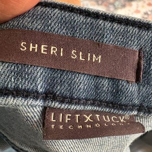 K&ouml;nnte beinhalten: Nahaufnahme einer Jeans aus Denim mit zwei braunen Etiketten. Auf dem oberen Etikett steht "SHERI SLIM" in wei&szlig;en Buchstaben. Auf dem unteren Etikett steht "LIFT X TUCK TECHNOLOGY" in wei&szlig;en Buchstaben.