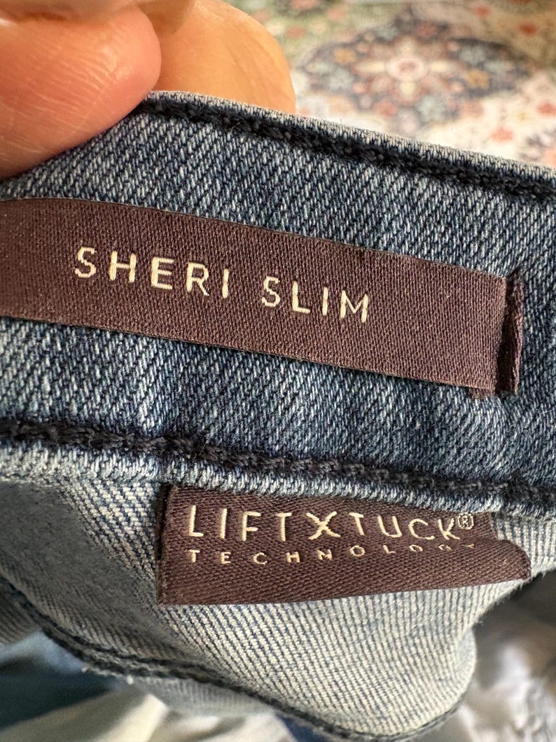 K&ouml;nnte beinhalten: Nahaufnahme einer Jeans aus Denim mit zwei braunen Etiketten. Auf dem oberen Etikett steht "SHERI SLIM" in wei&szlig;en Buchstaben. Auf dem unteren Etikett steht "LIFT X TUCK TECHNOLOGY" in wei&szlig;en Buchstaben.