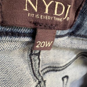 K&ouml;nnte beinhalten: Nahaufnahme eines braunen Etiketts mit dem Markennamen "NYDJ" und dem Satz "FIT IS EVERYTHING" in wei&szlig;er Schrift. Unter dem Markenschild befindet sich ein kleineres Etikett mit der Gr&ouml;&szlig;e "20W" in wei&szlig;er Schrift. Das Etikett ist an blauem Denim-Stoff befestigt.