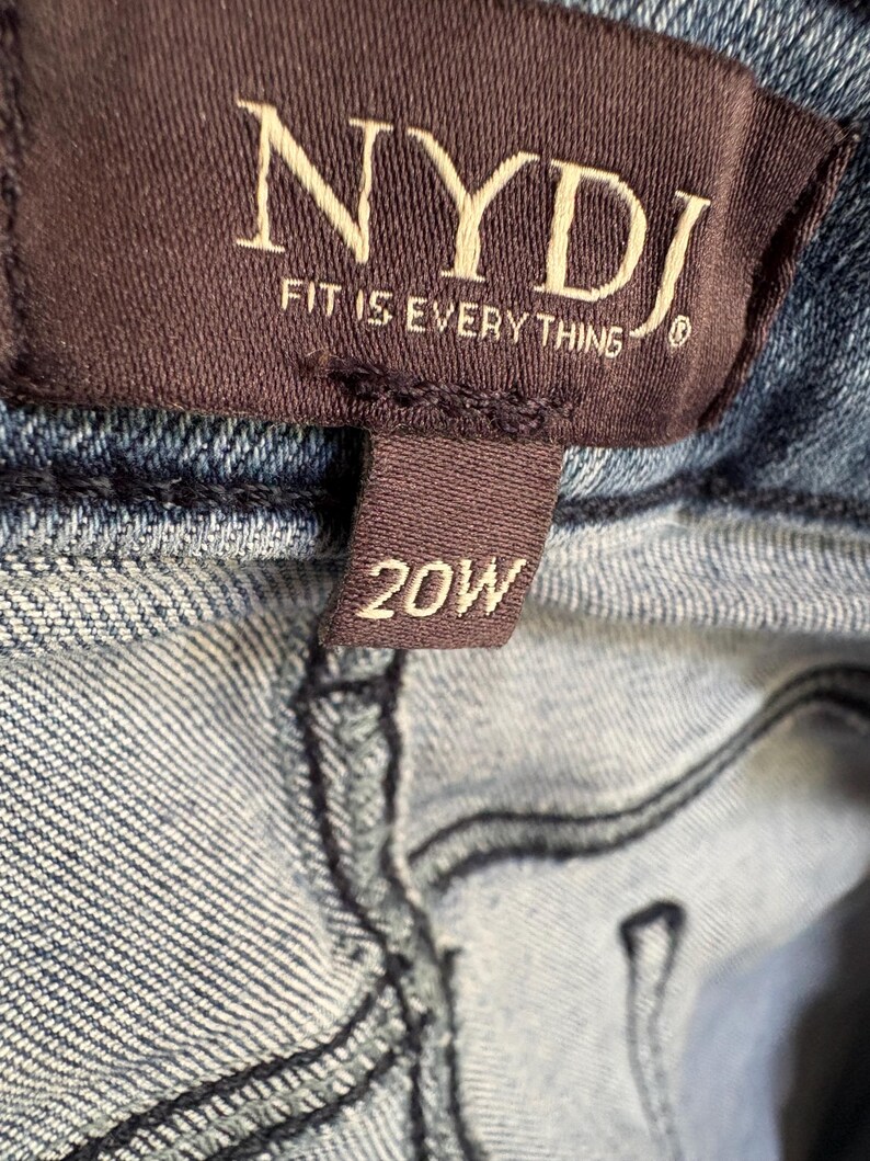 K&ouml;nnte beinhalten: Nahaufnahme eines braunen Etiketts mit dem Markennamen "NYDJ" und dem Satz "FIT IS EVERYTHING" in wei&szlig;er Schrift. Unter dem Markenschild befindet sich ein kleineres Etikett mit der Gr&ouml;&szlig;e "20W" in wei&szlig;er Schrift. Das Etikett ist an blauem Denim-Stoff befestigt.