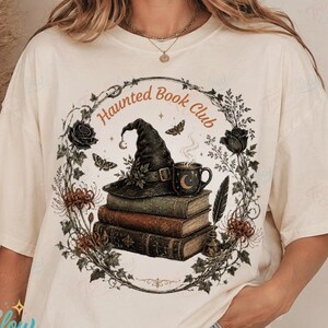 May include: A cream-coloured short-sleeve t-shirt featuring a graphic print of a stack of three books topped with a witch hat and a steaming mug. The design includes a circular floral wreath, moths, and the text Haunted Book Club in orange.