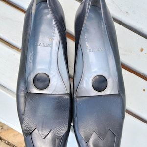 Può includere: Un paio di mocassini Bally in pelle nera. Le scarpe hanno una punta arrotondata con una patta decorativa e un accento circolare. Il logo Bally è visibile sulla soletta. Le scarpe sono esposte su una superficie chiara.