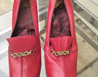 Könnte beinhalten: Ein Paar rote Leder-Loafer mit goldfarbenen Kettenakzenten. Die Schuhe haben eine spitze Zehenpartie und einen dekorativen Riemen über dem Oberteil. Das Innenfutter hat einen dunkelroten Musterstoff.