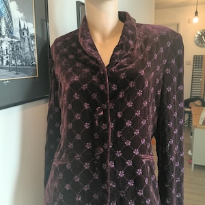 May include: A deep purple velvet blazer featuring a repeating diamond pattern with small floral accents at each intersection. The jacket has a shawl collar, long sleeves, and three fabric-covered buttons down the front centre for closure.