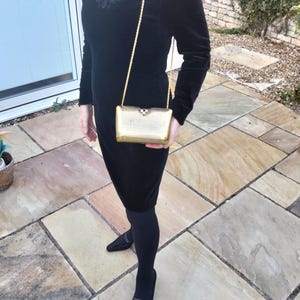 Puede incluir: Un vestido negro de terciopelo con mangas largas y escote en V, combinado con medias y zapatos negros. Se sostiene un bolso de mano rectangular dorado con una correa de cadena. El vestido tiene bordados decorativos alrededor del cuello.