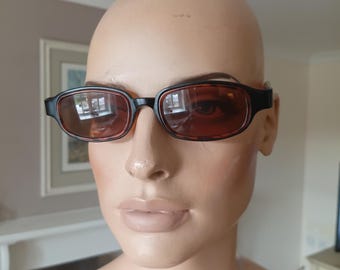 Puede incluir: Gafas de sol rectangulares negras y marrones con lentes tintadas. Las gafas de sol tienen un diseño clásico con montura oscura e interior marrón claro. Las lentes son de color ámbar cálido.