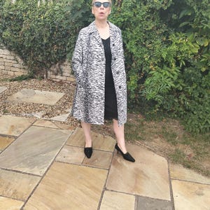 May include: A woman wearing a black dress, black heels, and sunglasses stands outdoors. She is wearing a long coat with a black and white zebra stripe pattern. The background includes greenery and stone paving.