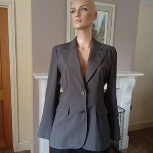 May include: A grey blazer with two buttons and two front pockets. The blazer is displayed on a mannequin. The blazer has long sleeves and a notched lapel. The mannequin is wearing a black skirt.