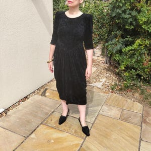 May include: A black velvet dress with three-quarter sleeves and a knee-length hem. The dress has a round neckline and a fitted bodice that flows into a pleated skirt. The person is wearing black sunglasses and black loafers.