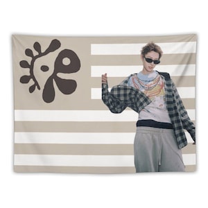 Puede incluir: Un tapiz a rayas beige y blanco con un diseño abstracto marrón oscuro en la esquina superior izquierda. Una persona con gafas de sol, una camiseta gráfica y una camisa a cuadros está de pie frente al tapiz.