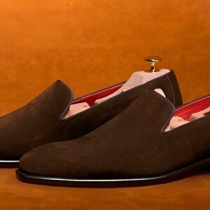 Pu&ograve; includere: Un paio di scarpe slip-on in camoscio marrone con tacco basso e suola nera. L'interno &egrave; foderato in tessuto rosso. Le forme sono inserite nelle scarpe. Le scarpe sono esposte su uno sfondo marrone.