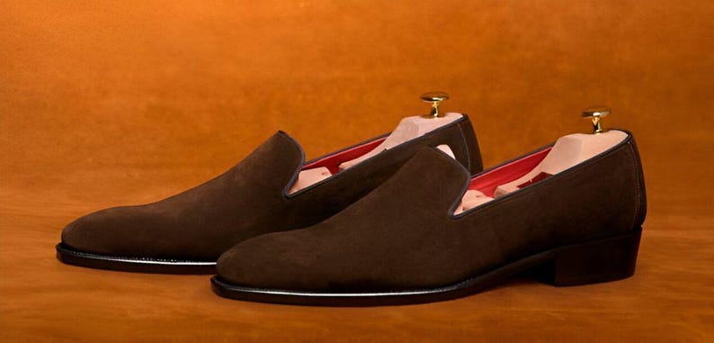 Pu&ograve; includere: Un paio di scarpe slip-on in camoscio marrone con tacco basso e suola nera. L'interno &egrave; foderato in tessuto rosso. Le forme sono inserite nelle scarpe. Le scarpe sono esposte su uno sfondo marrone.