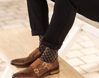 May include: Brown leather double monk strap shoes with suede accents and silver buckles. The shoes are paired with patterned socks and black trousers. The footwear is displayed on a wooden bench, showcasing a classic style.
