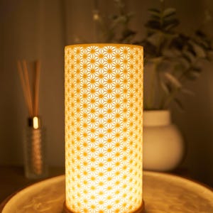 May include: A cylindrical lamp with a geometric star pattern, illuminated from within. The lamp sits on a wooden base and is placed on a decorative tray. The warm light creates a cosy ambiance.