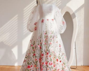 May include: A white, floor-length gown with floral embroidery. The dress features off-the-shoulder, sheer sleeves and a fitted bodice. The skirt is adorned with red, pink, and green floral patterns. The dress is displayed on a mannequin.