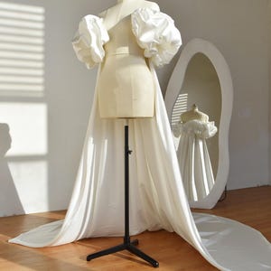 Puede incluir: Un vestido de novia color crema sobre un maniquí con mangas grandes y abullonadas. El vestido tiene una larga cola y se refleja en un espejo grande y de forma única. El maniquí está sobre un soporte negro, en una habitación luminosa.