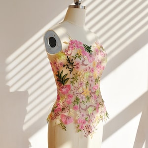 May include: A beige, floral-embroidered bodice on a mannequin. The bodice features pink, yellow, and green floral designs. The text "WattCustom" is visible on the bottom right. The garment is displayed in natural light.