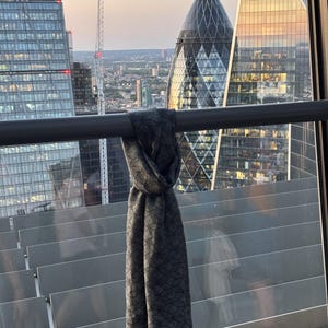 K&ouml;nnte beinhalten: Ein dunkelgrauer Schal mit geometrischem Muster h&auml;ngt vor dem Hintergrund moderner Wolkenkratzer. Der Schal ist &uuml;ber einem Metallgel&auml;nder drapiert, mit einer durch das Glas sichtbaren Stadtlandschaft. Die Textur und das Design des Schals sind deutlich sichtbar.