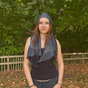 K&ouml;nnte beinhalten: Eine Person tr&auml;gt ein schwarzes Tanktop, einen dunkelgrauen Schal und ein passendes Kopftuch mit geometrischem Muster. Sie steht im Freien vor einem Holzzaun und gr&uuml;nem Laub. Der Schal ist um ihren Hals drapiert.