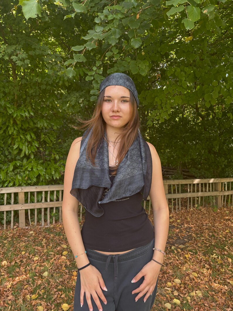 K&ouml;nnte beinhalten: Eine Person tr&auml;gt ein schwarzes Tanktop, einen dunkelgrauen Schal und ein passendes Kopftuch mit geometrischem Muster. Sie steht im Freien vor einem Holzzaun und gr&uuml;nem Laub. Der Schal ist um ihren Hals drapiert.
