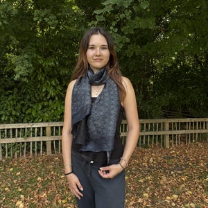 K&ouml;nnte beinhalten: Ein dunkelgrauer Schal mit geometrischem Muster wird um den Hals getragen. Der Schal besteht aus einem weichen Material und hat einen subtilen Glanz. Die Person tr&auml;gt ein schwarzes Tanktop und eine dunkelgraue Hose.
