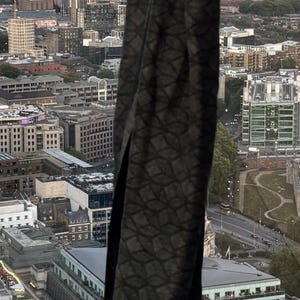 K&ouml;nnte beinhalten: Ein dunkelgrauer Schal mit geometrischem Muster wird vor einer Stadtkulisse gehalten. Das Design des Schals weist ein sich wiederholendes rautenf&ouml;rmiges Motiv auf. Der Hintergrund zeigt Geb&auml;ude und einen bew&ouml;lkten Himmel.