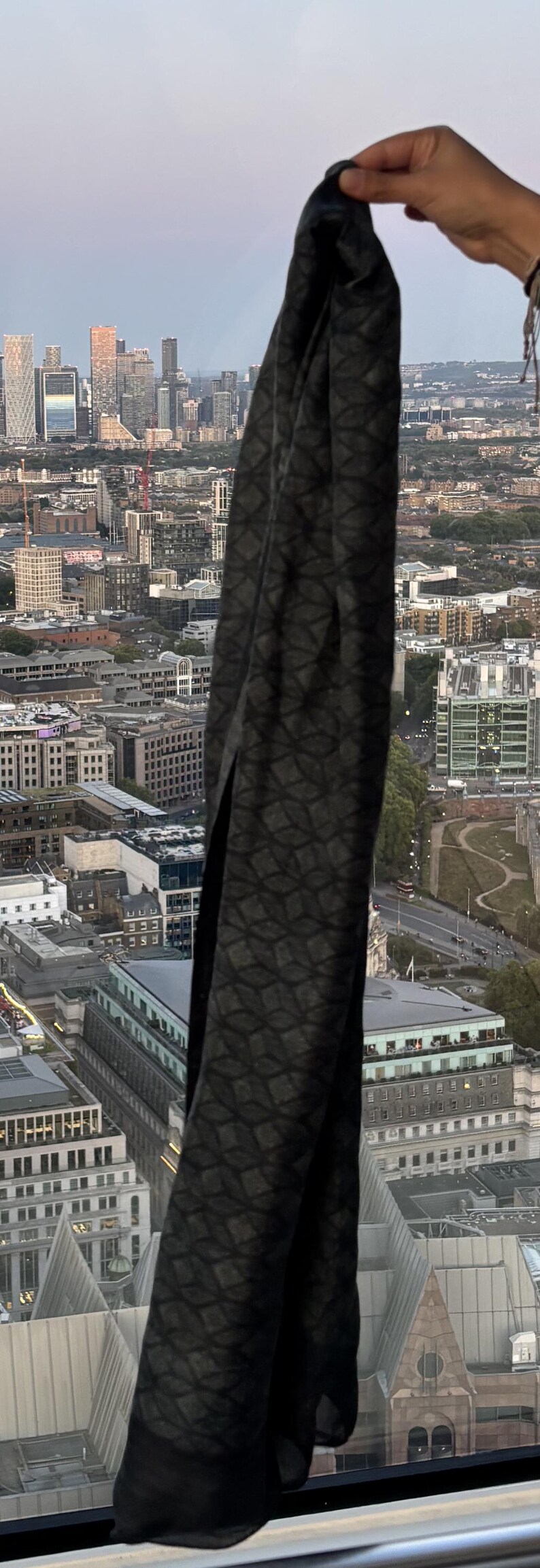 K&ouml;nnte beinhalten: Ein dunkelgrauer Schal mit geometrischem Muster wird vor einer Stadtkulisse gehalten. Das Design des Schals weist ein sich wiederholendes rautenf&ouml;rmiges Motiv auf. Der Hintergrund zeigt Geb&auml;ude und einen bew&ouml;lkten Himmel.