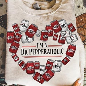 May include: A light beige sweatshirt with a heart-shaped arrangement of Dr Pepper soda cans. The cans are red and white, with the Dr Pepper logo. The text "I'M A DR PEPPERAHOLIC" is printed below the cans.