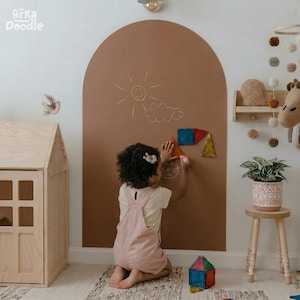 Puede incluir: Un niño con un mono rosa dibuja en una gran pizarra marrón en forma de arco. La pizarra tiene un dibujo de un sol y nubes. Formas magnéticas de colores están adheridas a la pizarra. Una casa de muñecas de madera y un móvil decorativo están en el fondo.