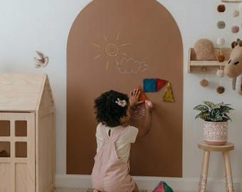May include: A large, arch-shaped brown wall decal featuring a hand-drawn sun and cloud. The decal serves as a creative space for play, with colorful translucent geometric magnetic tiles being placed on the surface by a child in pink overalls.