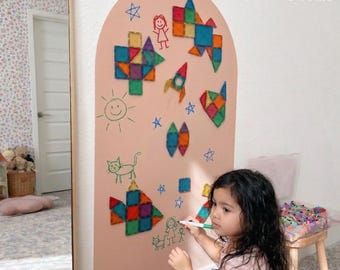 May include: An arched, light pink magnetic wall board featuring colorful geometric magnetic tiles arranged into shapes like a rocket, fish, and abstract patterns, alongside hand-drawn green and red marker illustrations of people, a cat, and stars.