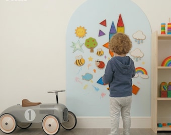 May include: A light blue arched wall decal featuring colorful magnetic shapes including a sun, tree, bees, ladybug, clouds, and a rainbow. The Arka Doodle branding is visible in the top left corner. A grey ride-on toy car sits on the floor nearby.