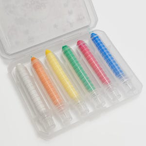 May include: A clear plastic case containing six colorful crayons. The crayons are white, orange, yellow, green, red, and blue. Each crayon has a clear plastic casing and a white tip. The case is open, revealing the crayons inside.