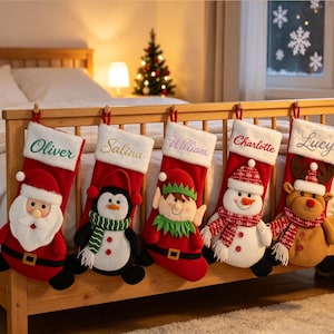 Può includere: Cinque calze natalizie rosse e bianche appese a una struttura letto in legno. Ogni calza presenta un personaggio in peluche: Babbo Natale, un pinguino, un elfo, un pupazzo di neve e una renna. I risvolti bianchi sono ricamati con i nomi Oliver, Salina, William, Charlotte e Lucy.
