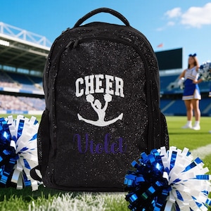 Puede incluir: Una mochila negra con purpurina que presenta el texto bordado en blanco CHEER sobre una silueta blanca de una animadora realizando un salto. El nombre Violet está bordado en letra cursiva morada debajo del gráfico. Tiene cremalleras negras.