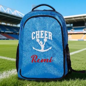 Peut inclure: Un sac à dos bleu vif à paillettes orné d'un motif blanc représentant une personne faisant un saut de cheerleading avec le texte CHEER au-dessus et le prénom Remi en lettres rouges. Le sac possède des bordures noires et des poches latérales.