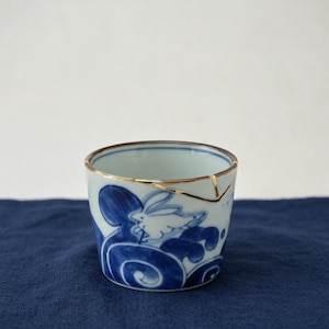 May include: A small ceramic cup featuring a white rabbit illustration amidst blue swirling wave patterns. The rim of the cup displays a distinct gold-colored repair line, indicating a kintsugi technique used to mend a crack in the porcelain surface.