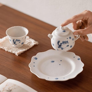 Op de afbeelding: Een witte keramische theeset met blauwe kattenillustraties. De set bevat een kleine theepot, een theekopje en een bord met een geschulpte rand. De theepot wordt door een hand vastgehouden. Het theekopje staat op een kleine, vierkante, beige onderzetter.
