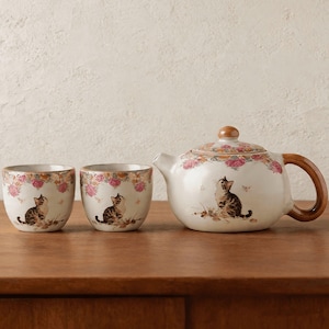 Op de afbeelding: Een keramische theeset met een theepot en twee kopjes. De set is versierd met een bloemenpatroon en een illustratie van een tabby kat. De theepot heeft een bruin handvat en deksel. De set staat op een houten oppervlak.