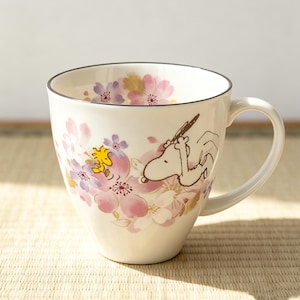 May include: A white ceramic mug featuring a delicate watercolour-style illustration of Snoopy and Woodstock surrounded by pink and purple cherry blossoms. The mug has a smooth, tapered body, a curved handle, and a thin dark rim along the top edge.