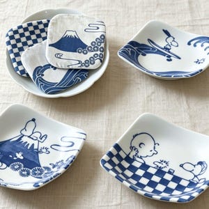 Könnte beinhalten: Ein Set aus drei quadratischen weißen Keramikschalen und einer weißen Schüssel mit Stoffuntersetzern. Die Schalen sind mit blauen Illustrationen von Snoopy, Charlie Brown, dem Berg Fuji und Karomustern verziert. Die Untersetzer zeigen ebenfalls blaue Designs.