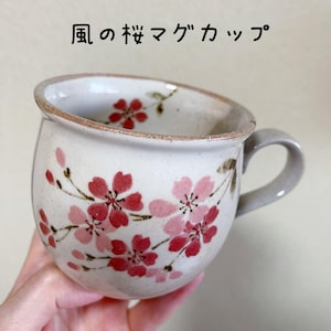 May include: A ceramic mug with a speckled off-white glaze featuring hand-painted pink and red cherry blossom motifs on the exterior and interior rim. The text 風の桜マグカップ is printed above the mug. It has a rounded body and a curved handle.